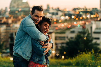 Romantic diverse couple embracing and looking away while standing on lawn against cityscape with buildings on blurred background