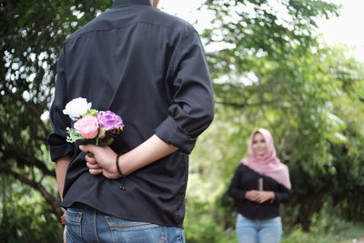 Rear view of boyfriend holding flowers with girlfriend in background at park