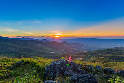 Woman sitting on rock against sky during sunset