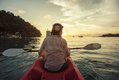 Rear view of young woman sitting on boat in sea against sky during sunset