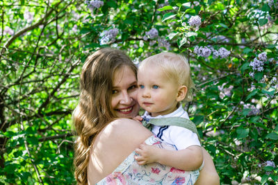 Candid portrait of young smiling mother with her little son in garden with blooming lilac flowers.