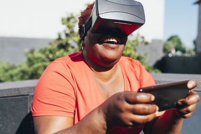 Playful woman wearing virtual reality holding smart phone on sunny day