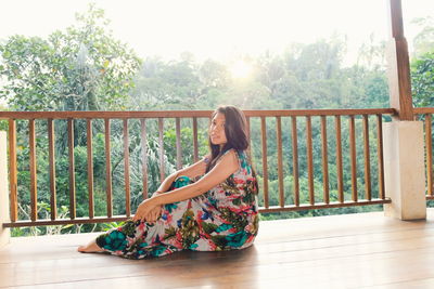 Portrait of young woman in dress sitting on hardwood floor by railing against sky