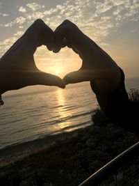 Hand holding heart shape in sea against sky during sunset