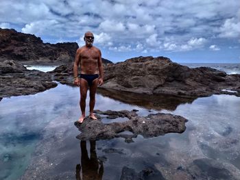 Full length of shirtless man standing on rock at sea