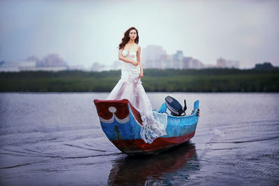 Beautiful female model in white evening gown standing on boat on shore