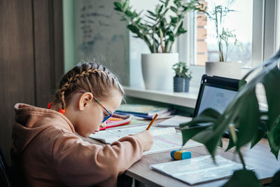 Primary school girl in eyeglasses writing in her notebook while having online lesson during covid