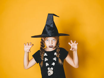 Portrait of girl wearing witch hat gesturing against yellow background during halloween