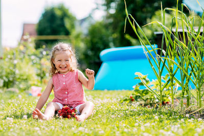 A charming little girl is sitting on the lawn in the garden, eating strawberries and laughing. 