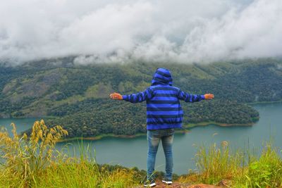 Rear view of man with arms outstretched standing on rock by lake