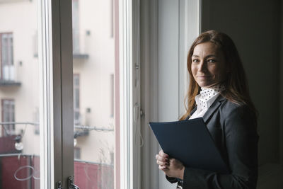 Portrait of confident mature female real estate agent standing with brochure by window at home