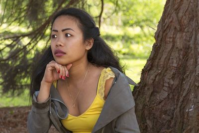 Young woman looking away while sitting against tree trunk