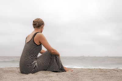 Woman sitting at sandy beach against cloudy sky