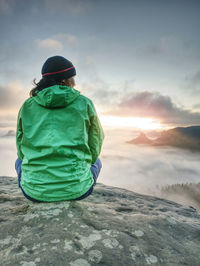 Green jacket woman pensively sitting on the edge of a rock and looking at the misty clouds.