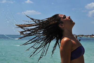 Side view of woman tossing wet hair by sea against sky