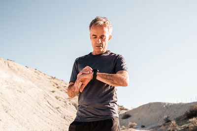 Concentrated middle aged male athlete in activewear checking time on smartwatch while standing in hilly terrain after workout