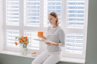 Woman reading book while sitting by window at home