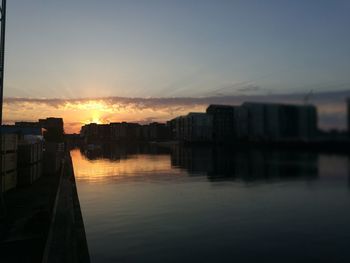 Silhouette buildings by river against sky during sunset