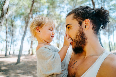 Side view of loving ethnic father gently cuddling cute child in cheek while standing in woods together at weekend