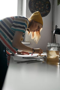 Side view of delighted female artist in trendy wear creating artwork in studio while painting on paper with watercolors