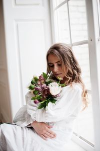 Bride holding bouquet while sitting at window sill in room