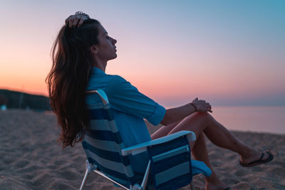 Woman looking at while sitting on beach against sky during sunset