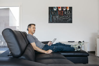 Mature man with digital tablet looking away while sitting on sofa at home