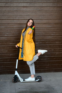 A girl in a yellow raincoat stands on a white scooter and holds colorful ice cream in her hands