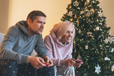 Fun young family in home clothes playing video games on couch on background of christmas tree