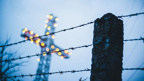Low angle view of barbed wire fence and illuminated cross against sky at dusk