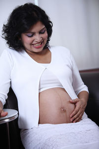 Smiling pregnant woman touching belly while sitting on sofa at home