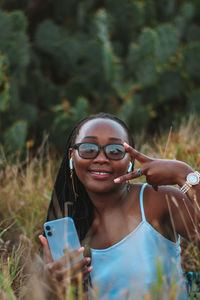 Portrait of young woman wearing sunglasses while holding  a phone showing a peace sign