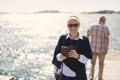 Portrait of smiling senior woman wearing sunglasses while holding mobile phone against lake