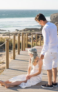 Full length of woman sitting by man standing on pier against sky