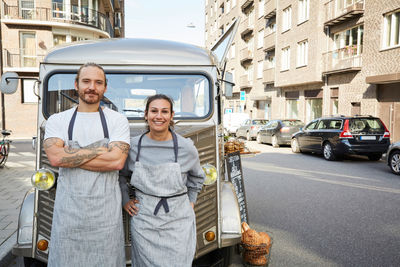 Portrait of confident male and female owners standing in front of food truck parked on city street