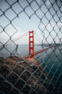 Golden gate bridge over bay against sky seen through damaged chainlink fence