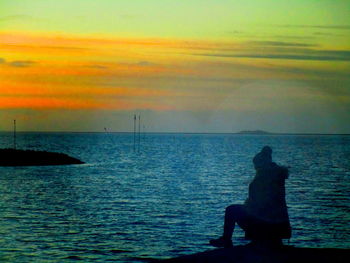 Silhouette man sitting by sea against sky during sunset