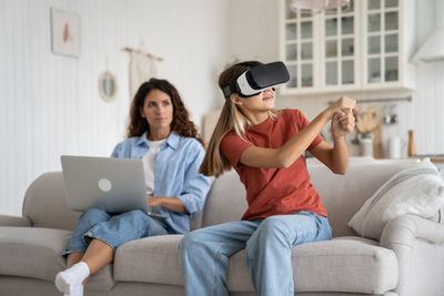 Enthusiastic positive teenage girl with vr glasses with virtual baseball bat sits on couch near mom