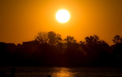 Silhouette trees by lake against orange sky during sunset