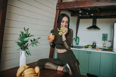 Smiling woman holding juice eating grapes at home