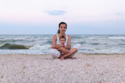 Portrait of mother and daughter sitting on shore at beach against sky