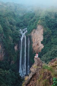 Man sitting on cliff against waterfall in forest