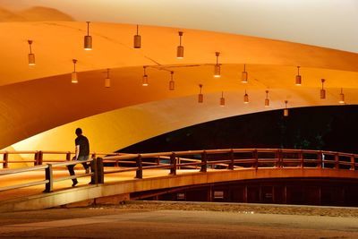 Full length of man sitting on railing below illuminated orange bridge at night