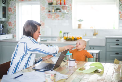 Multi-tasking female freelancer feeding food to daughter while telecommuting at dining table