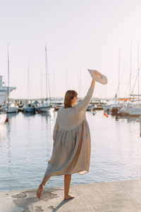 Happy young woman in dress and hat walks on the beach near the boats