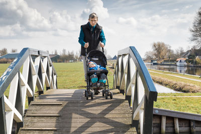Grandfather pushing granddaughter sleeping in baby stroller on footbridge against sky