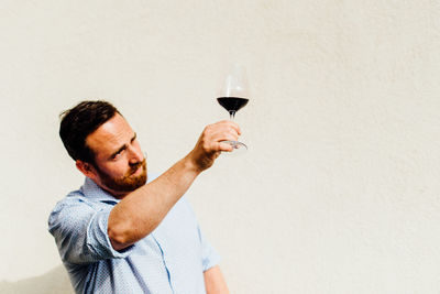 A man looking at glass of red wine while standing against white wall