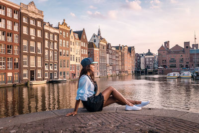 Woman sitting by canal in city against sky