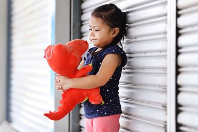 Girl holding stuffed toy while standing by shutter