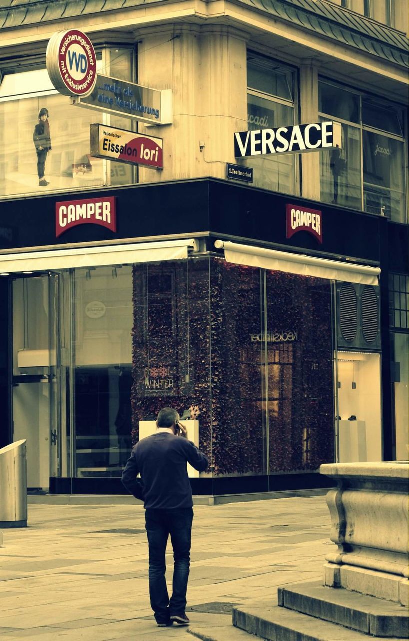 REAR VIEW OF MAN WALKING ON FOOTPATH IN FRONT OF BUILDINGS